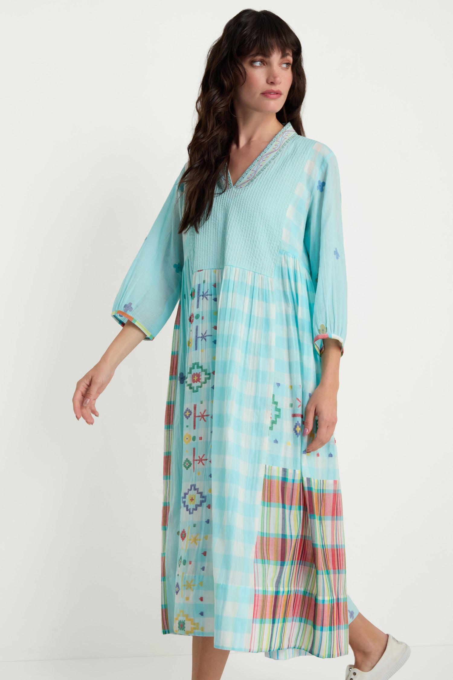 A woman models the Pintuck Jamdani Dress, featuring light blue cotton fabric with geometric and plaid woven motifs and a pintuck front panel, set against a plain white background.
