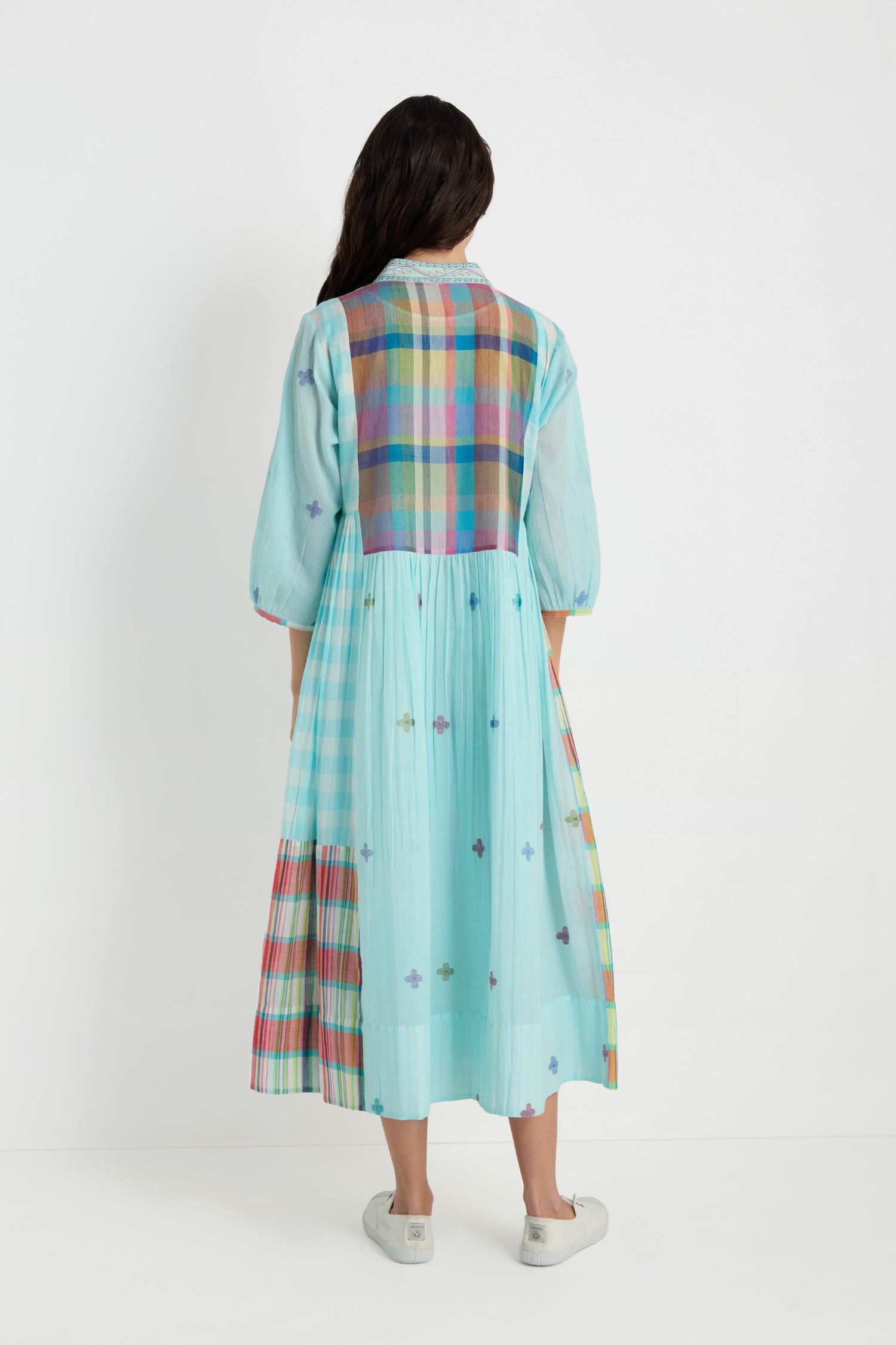 A person stands facing away, wearing the Pintuck Jamdani Dress—light blue cotton with multicolored plaid, woven motifs, and a pintuck front panel—paired with white sneakers against a plain white background.