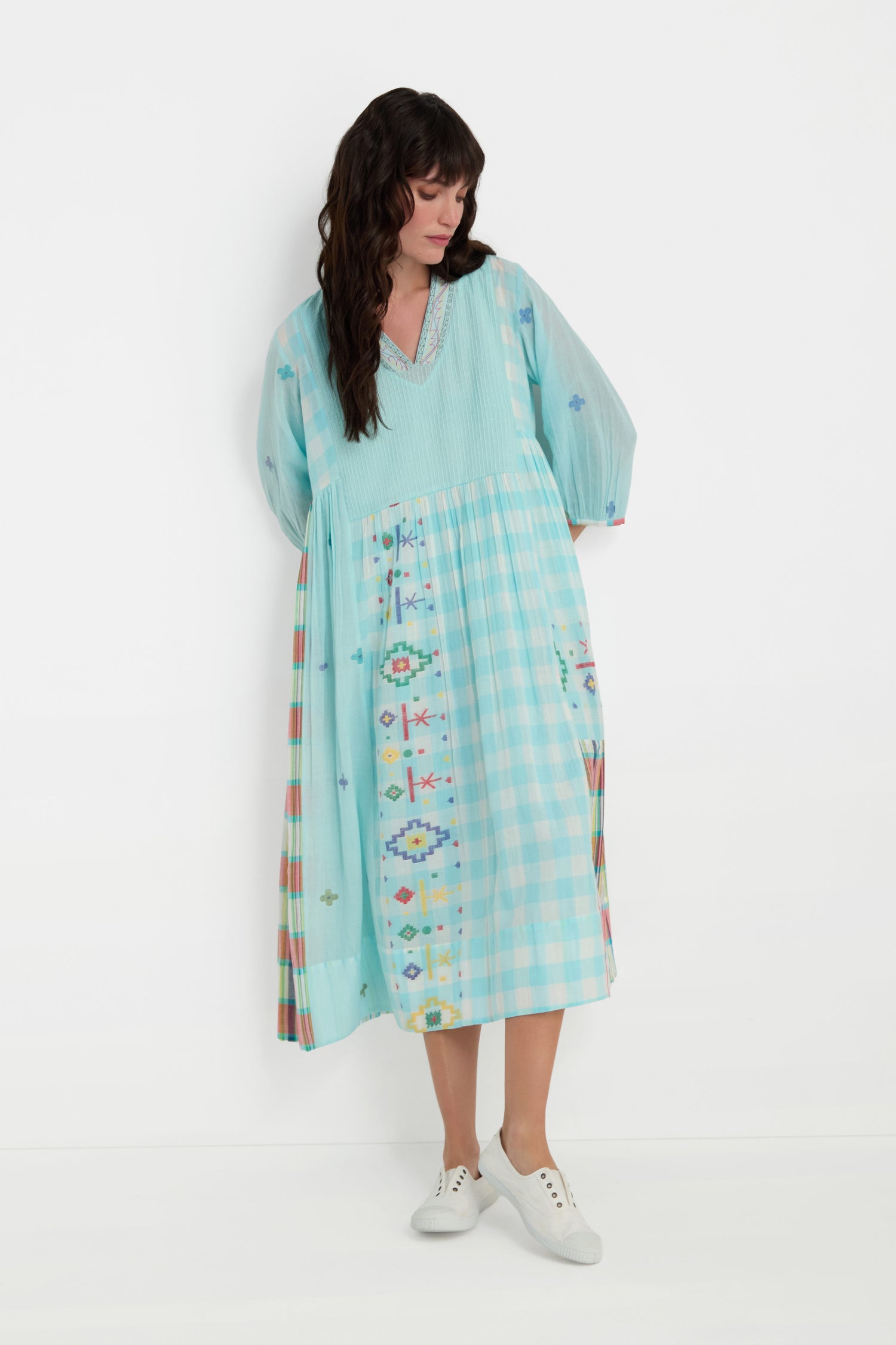 A woman wears the Pintuck Jamdani Dress, a loose-fitting, light blue cotton dress with multicolored woven motifs, and white sneakers. She stands against a plain white background, looking down.