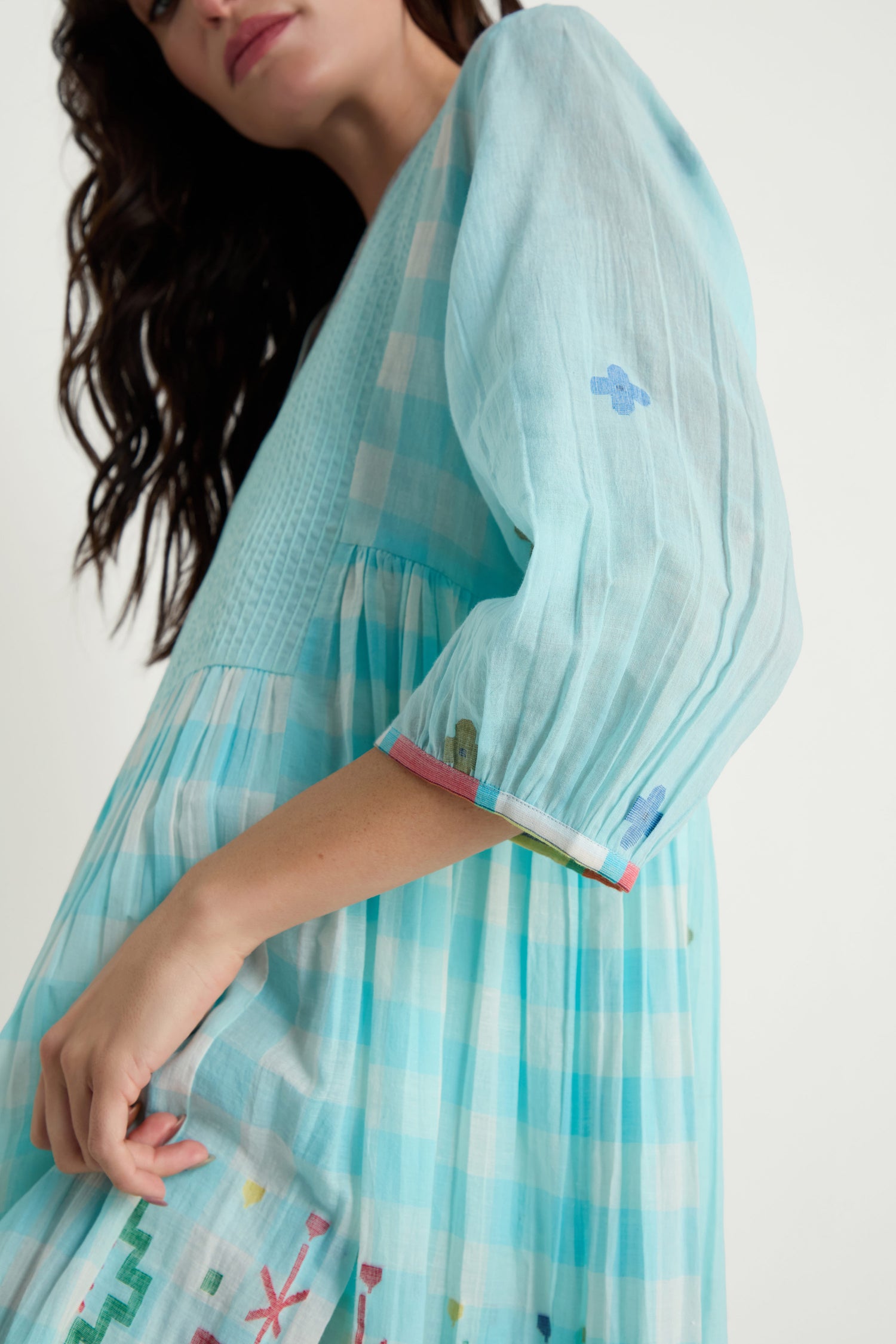 A woman models the Pintuck Jamdani Dress, featuring a light blue and white checkered cotton fabric with woven motifs, embroidery, pintuck front panel, and puffed sleeves against a plain backdrop.