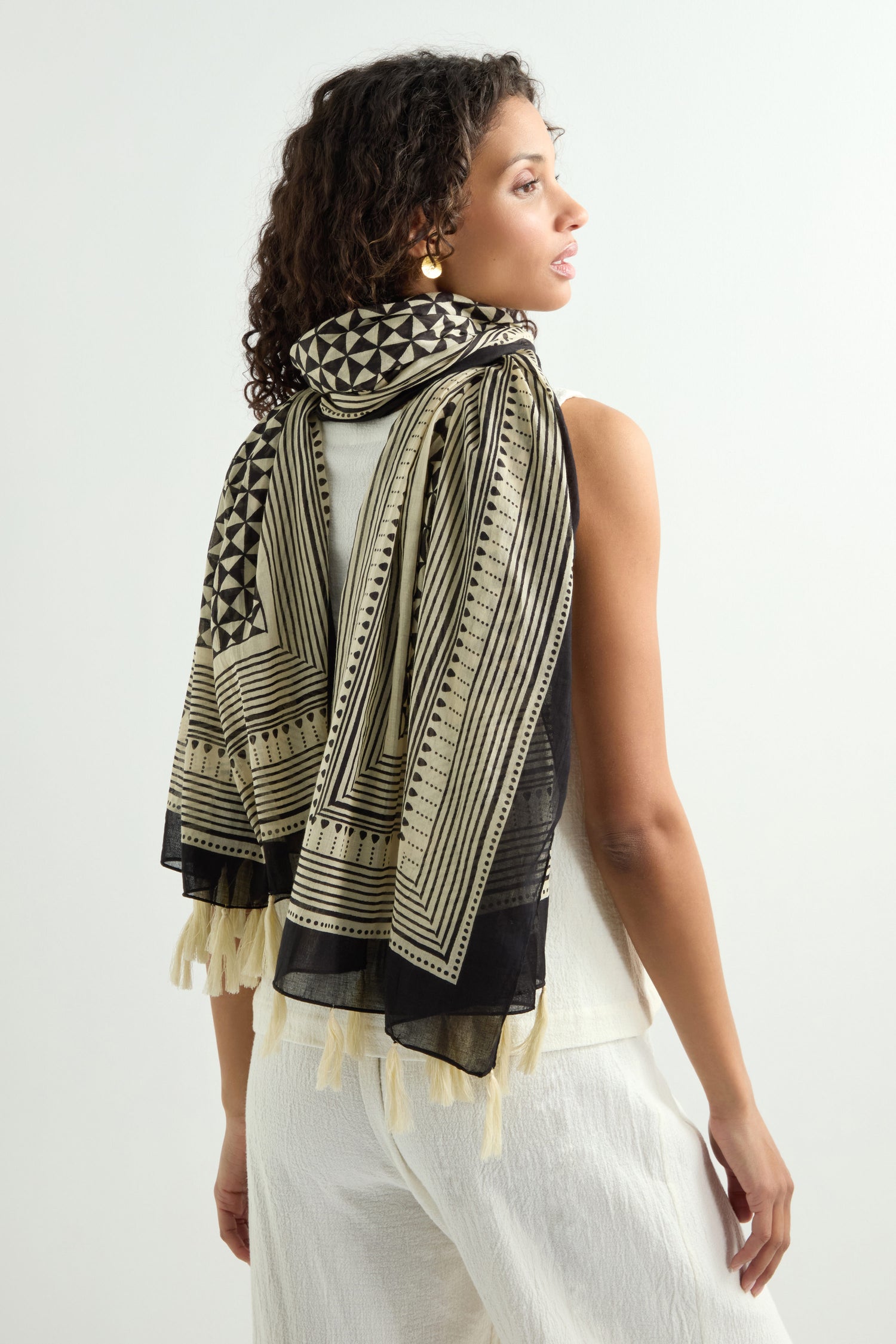 A woman in a sleeveless white outfit and gold earrings poses with her back turned, elegantly draping the Tassel Cotton Scarf—featuring a hand-printed black and beige pattern with tassel trim—over her shoulders.