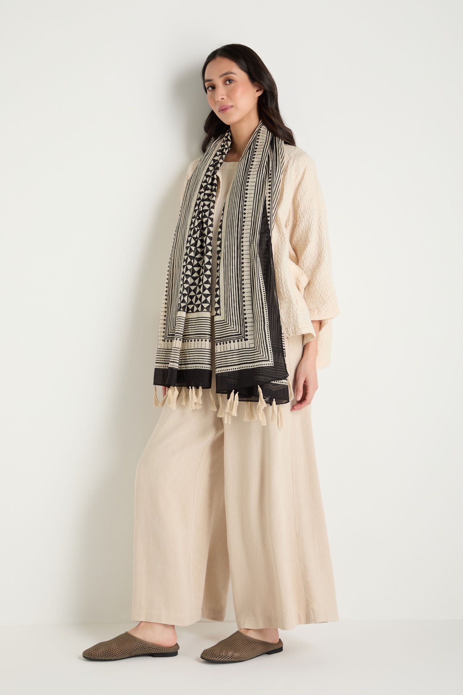 A woman stands against a white background wearing a loose cream outfit with wide-leg pants, a textured top, brown slip-on shoes, and the Cotton Tassel Scarf.