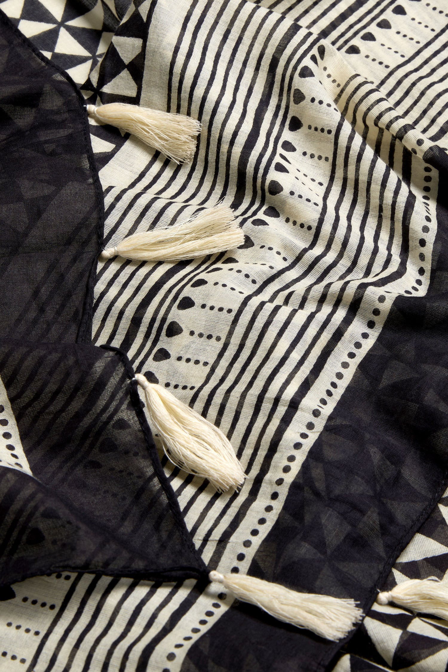 A close-up of the Cotton Tassel Scarf shows its geometric black patterns on a beige background, finished with white tassels along the edge.