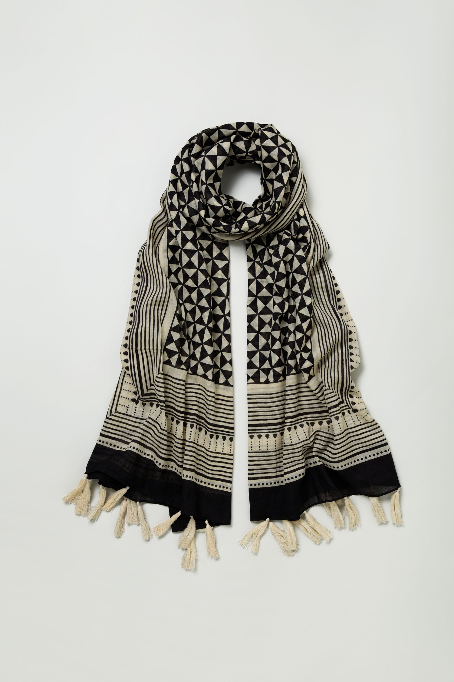 The Cotton Tassel Scarf features a black and beige geometric print with stripes and a striped border, plus fringed cotton tassels at both ends, shown laid flat on a light background.