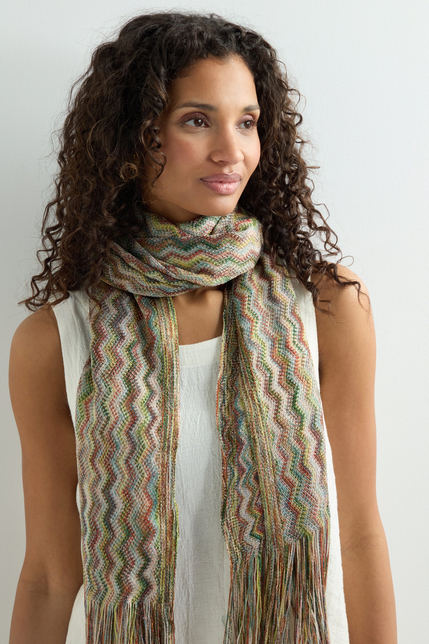 A woman with curly hair stands against a plain light background, wearing a sleeveless white top and the Zig Zag Fringe Viscose Scarf, which features a vibrant multi-coloured zigzag pattern and fringed edges for bohemian flair.