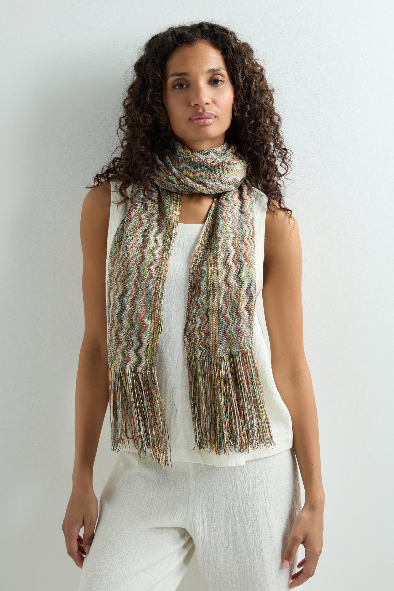 A woman with curly hair exudes boho style, wearing the Zig Zag Fringe Viscose Scarf, which features a multi-coloured zigzag pattern, draped over a white sleeveless top and pants against a simple light backdrop.
