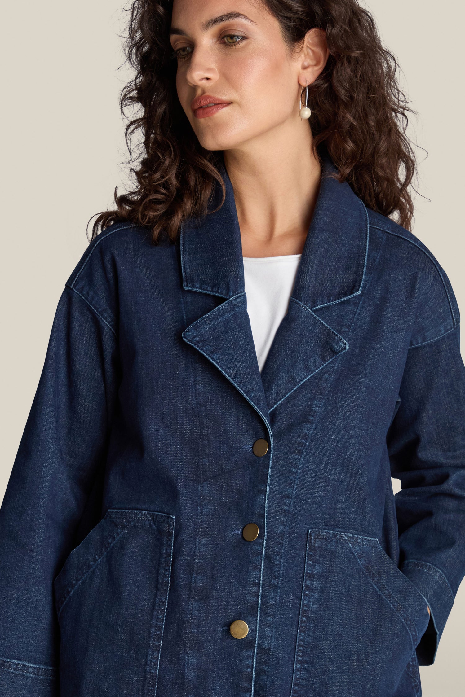 A woman with curly hair wearing the Stretch Denim Jacket over a white top, featuring a relaxed silhouette, looking to the side.