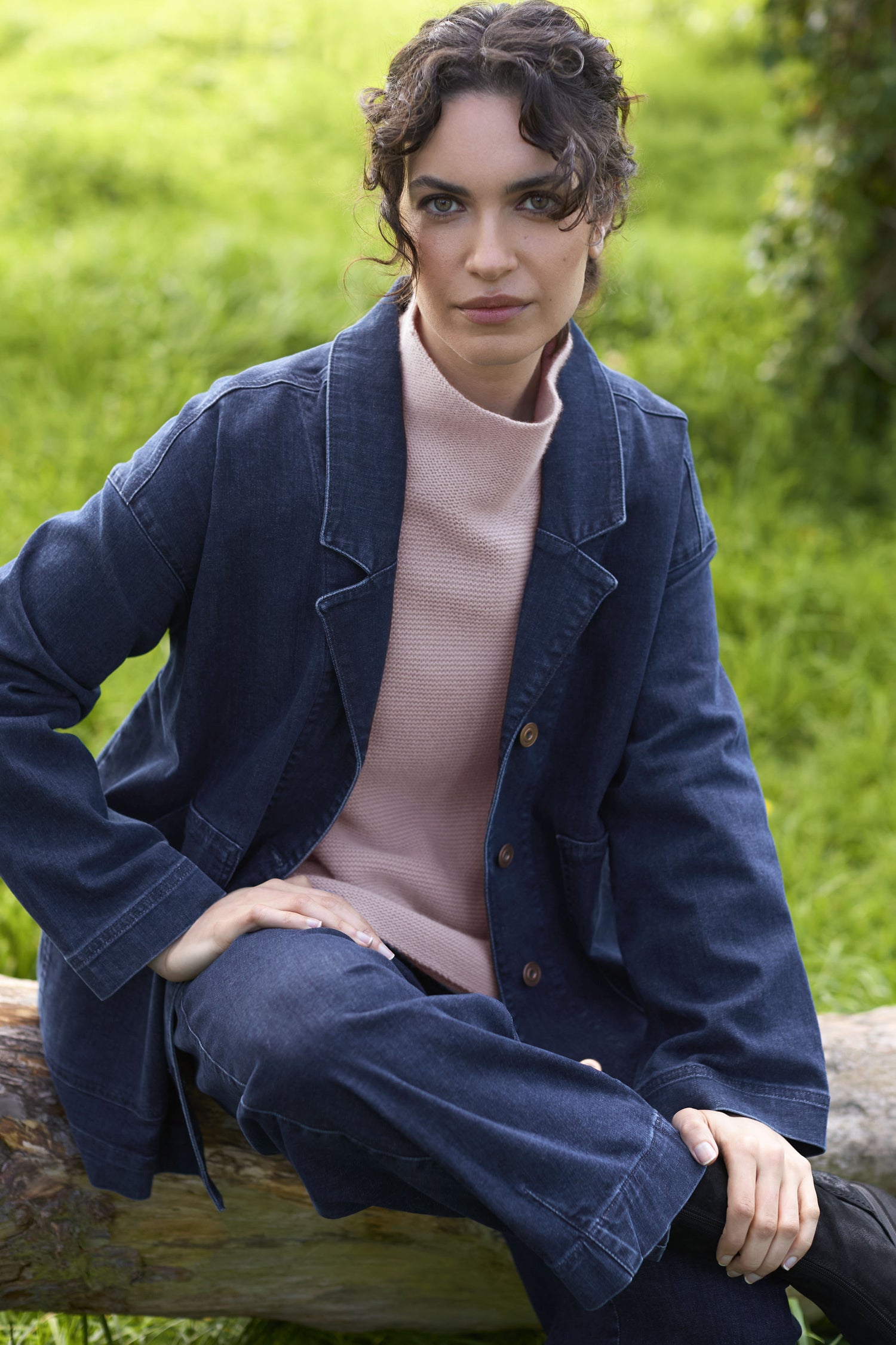 A woman sits on a log outdoors, wearing a Stretch Denim Jacket and matching pants paired with a light-colored top. Her relaxed silhouette blends seamlessly with the natural backdrop of green grass and foliage as she gazes at the camera with a neutral expression.