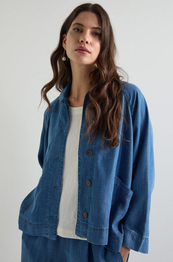 A woman wearing the Fluid Denim Boxy Jacket over a white top gazes into the camera. Her long brown hair contrasts with the plain white background, accentuating her modern style.