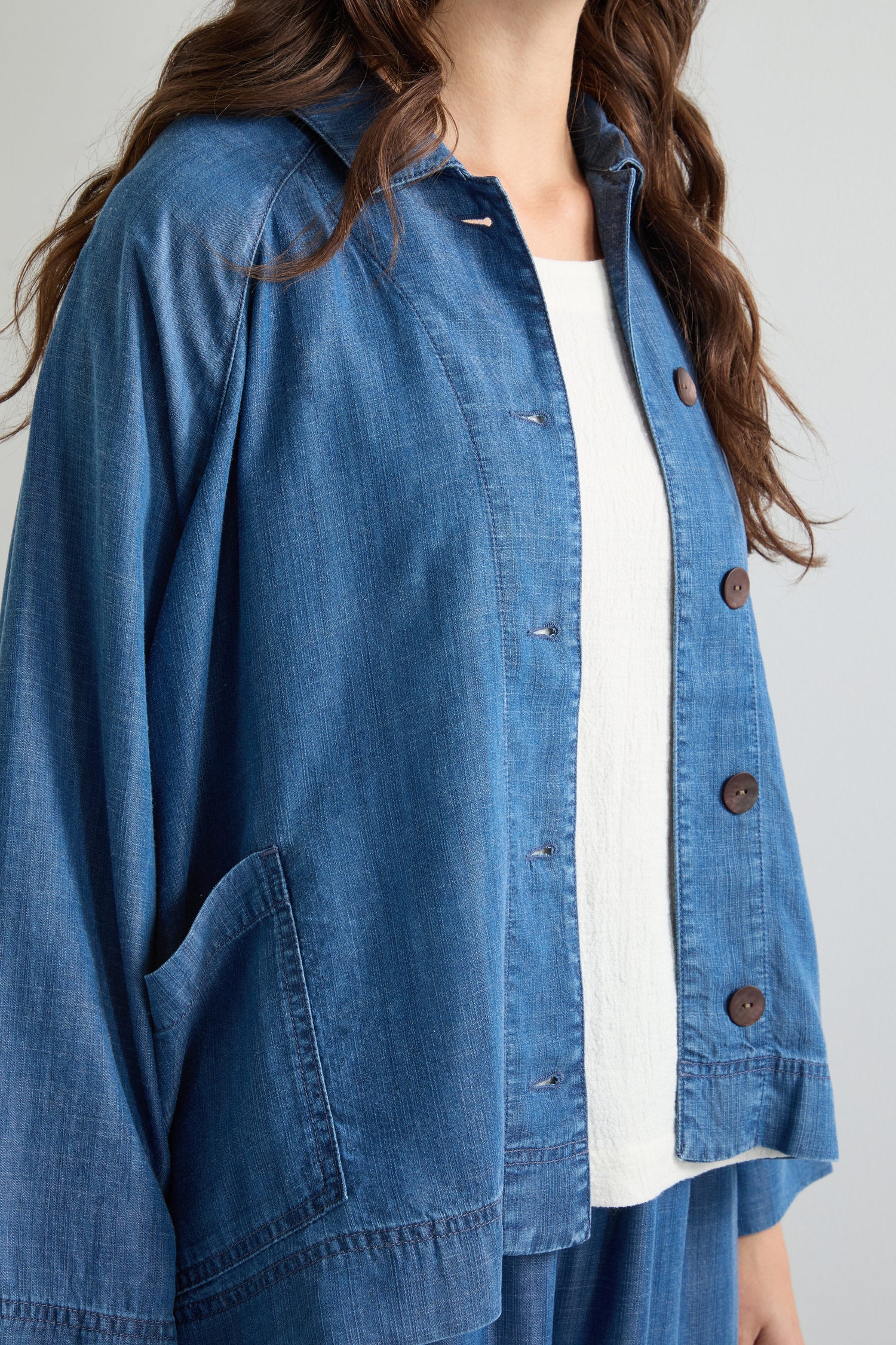 Wearing a Fluid Denim Boxy Jacket over a white top, one sports an effortlessly chic look. This blue denim jacket is characterized by its buttoned design and front pocket.