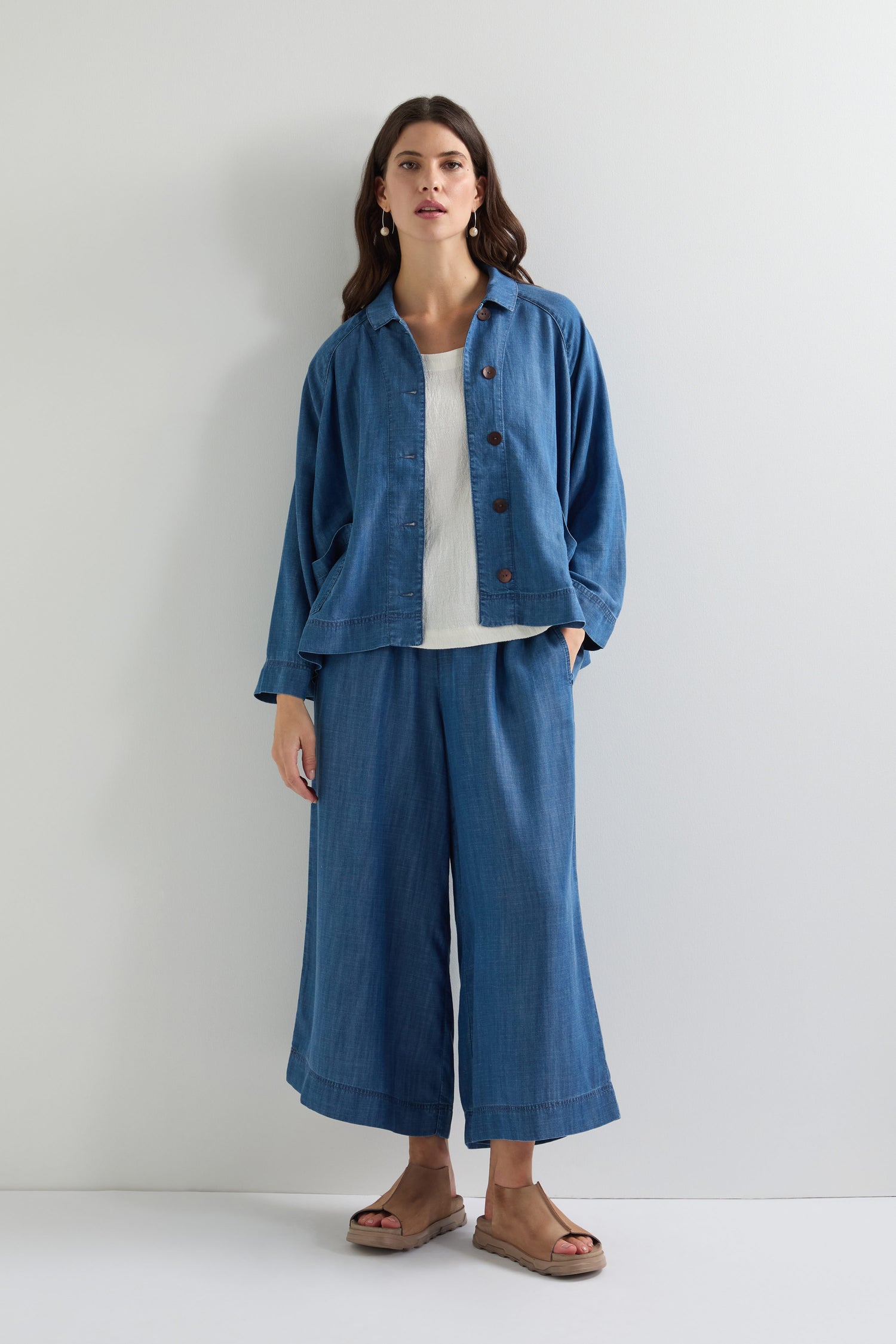 A woman poses against a simple backdrop in a Fluid Denim Boxy Jacket, wide-leg pants, white shirt, and sandals.