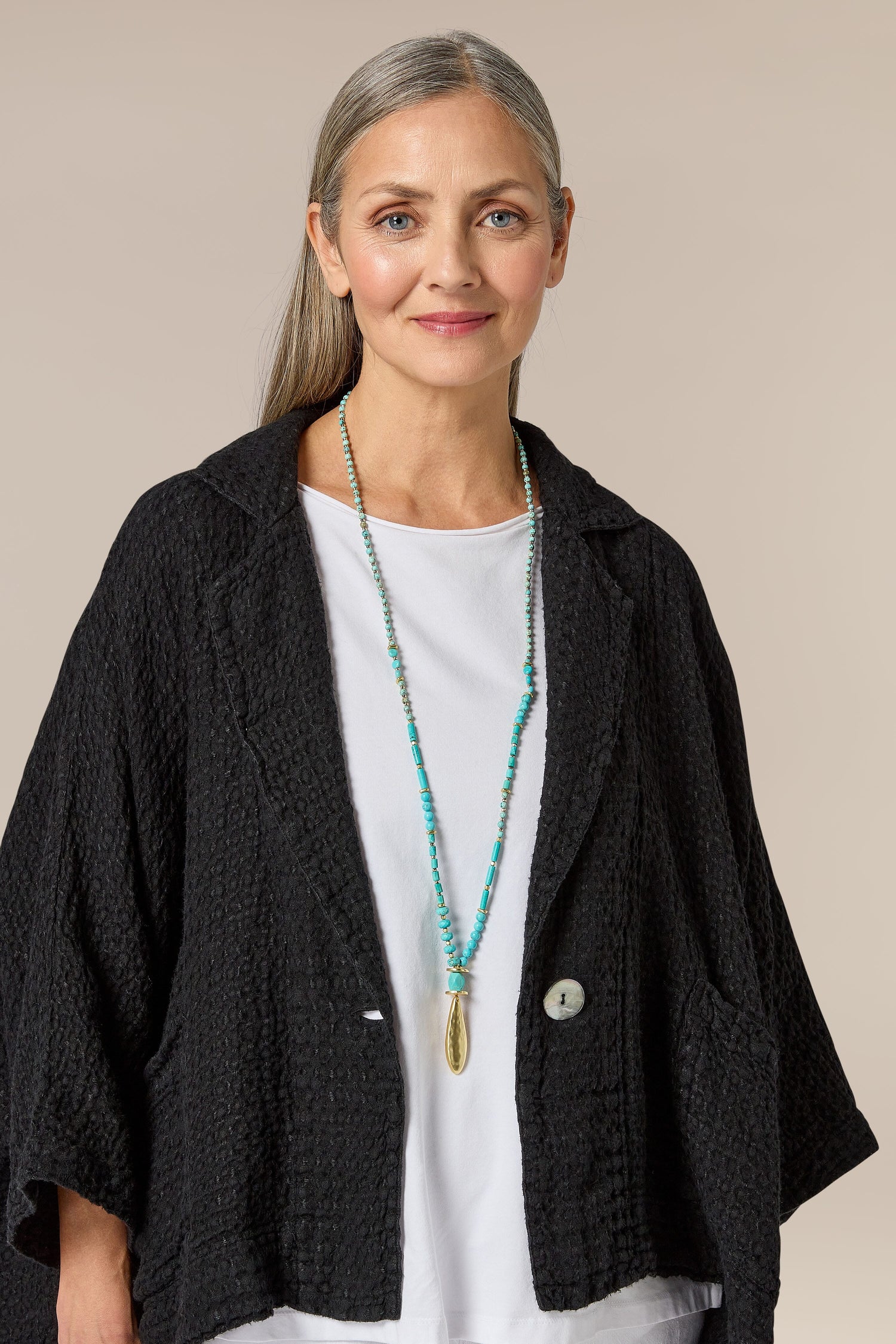 A woman donning a handcrafted black jacket, adorned with a turquoise necklace featuring a Bohemian accessory and a stunning Turquoise Bead Gold Pendant.