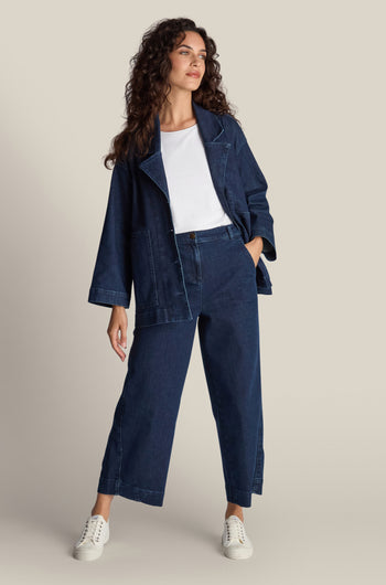 A woman with long, wavy hair stands wearing a dark denim jacket, matching high waist Stretch Denim Barrel Leg Trousers, a white shirt, and white sneakers. She has her left hand in her pocket and looks slightly to the side.