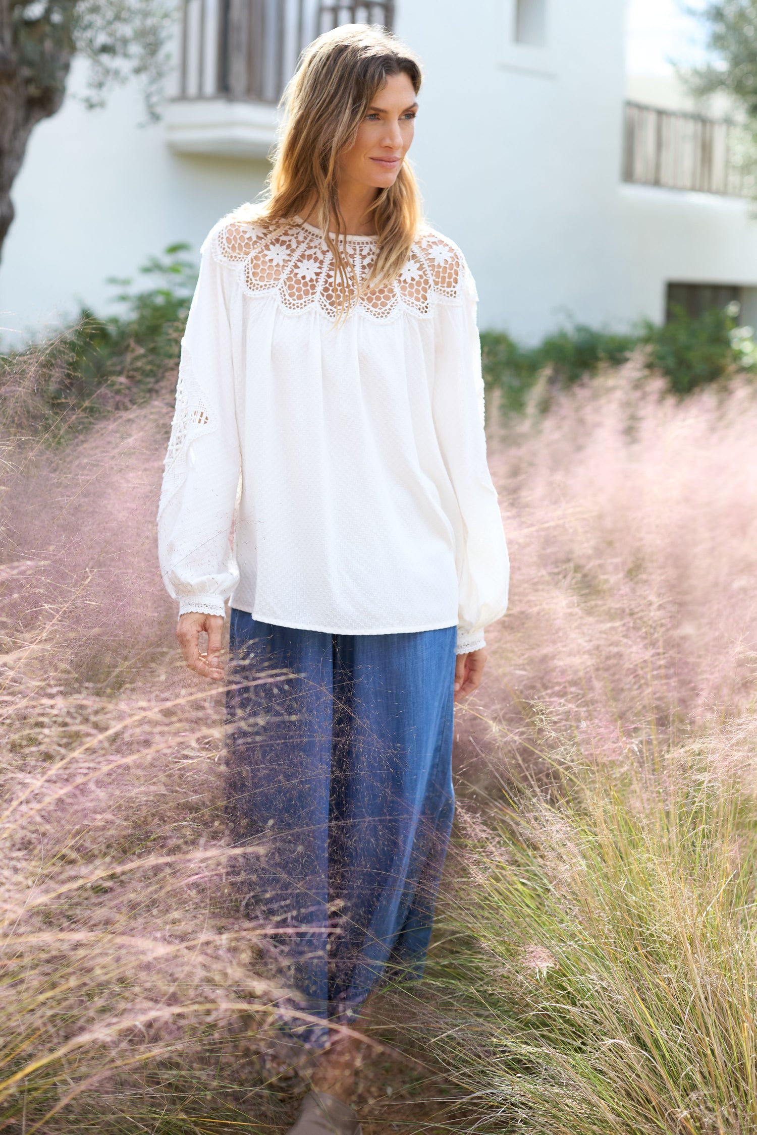 A woman wearing a white blouse and Fluid Denim Wide Trouser stands gracefully in a field of tall grass, with a white building softly visible in the background.