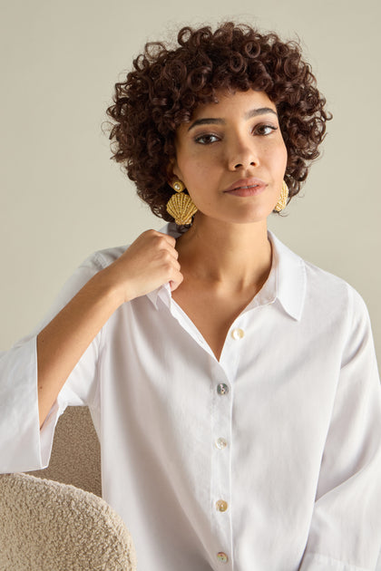 A person with curly hair, wearing a white shirt and Hand Embroidered Golden Shell Glass Pearl Earrings, sits gracefully on a textured chair.