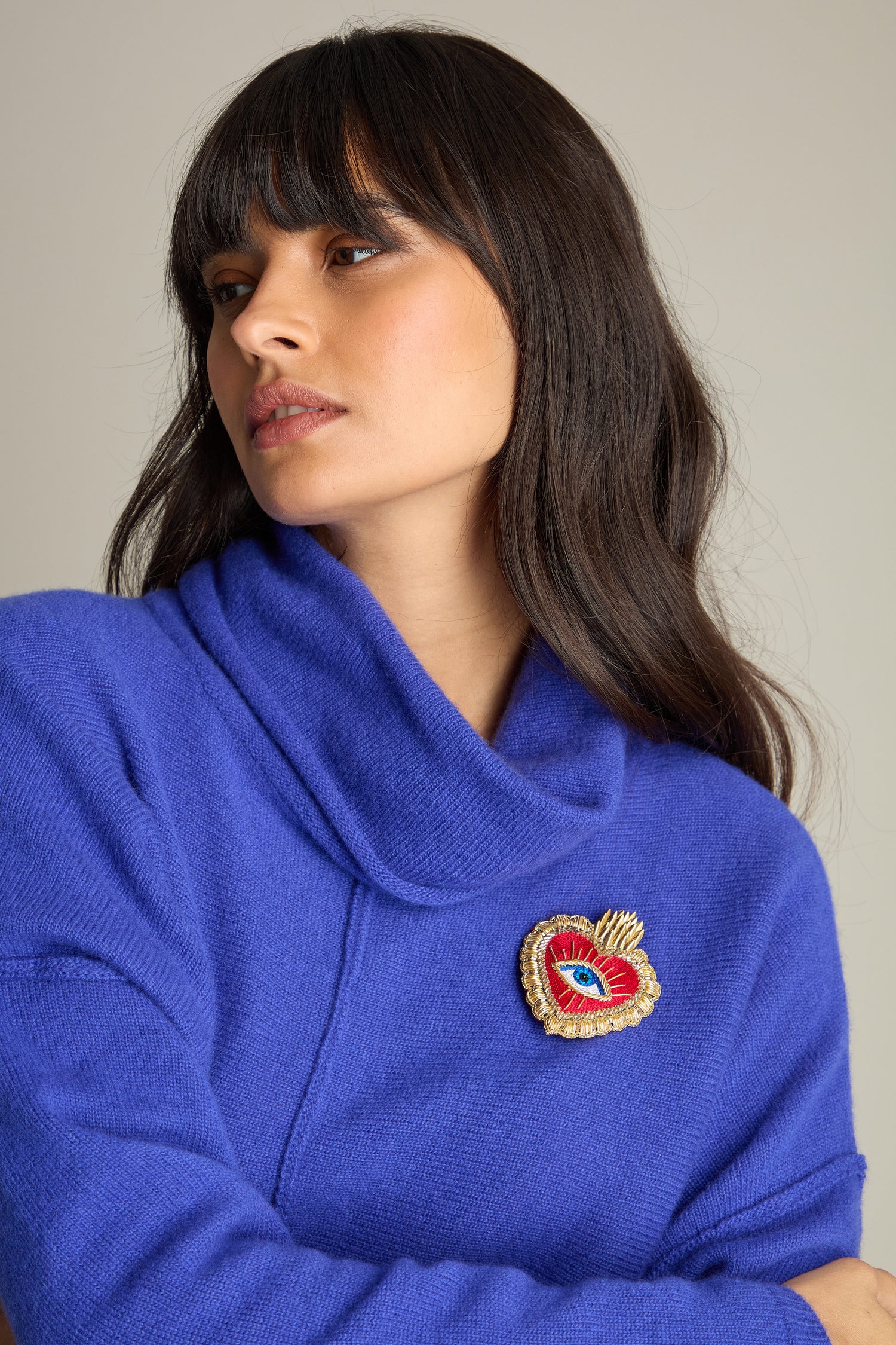 A person with long, dark hair and bangs wears a royal blue sweater, adorned with a Hand Beaded Dali Heart Brooch featuring an eye. The wearable art piece adds a unique touch against the plain background.