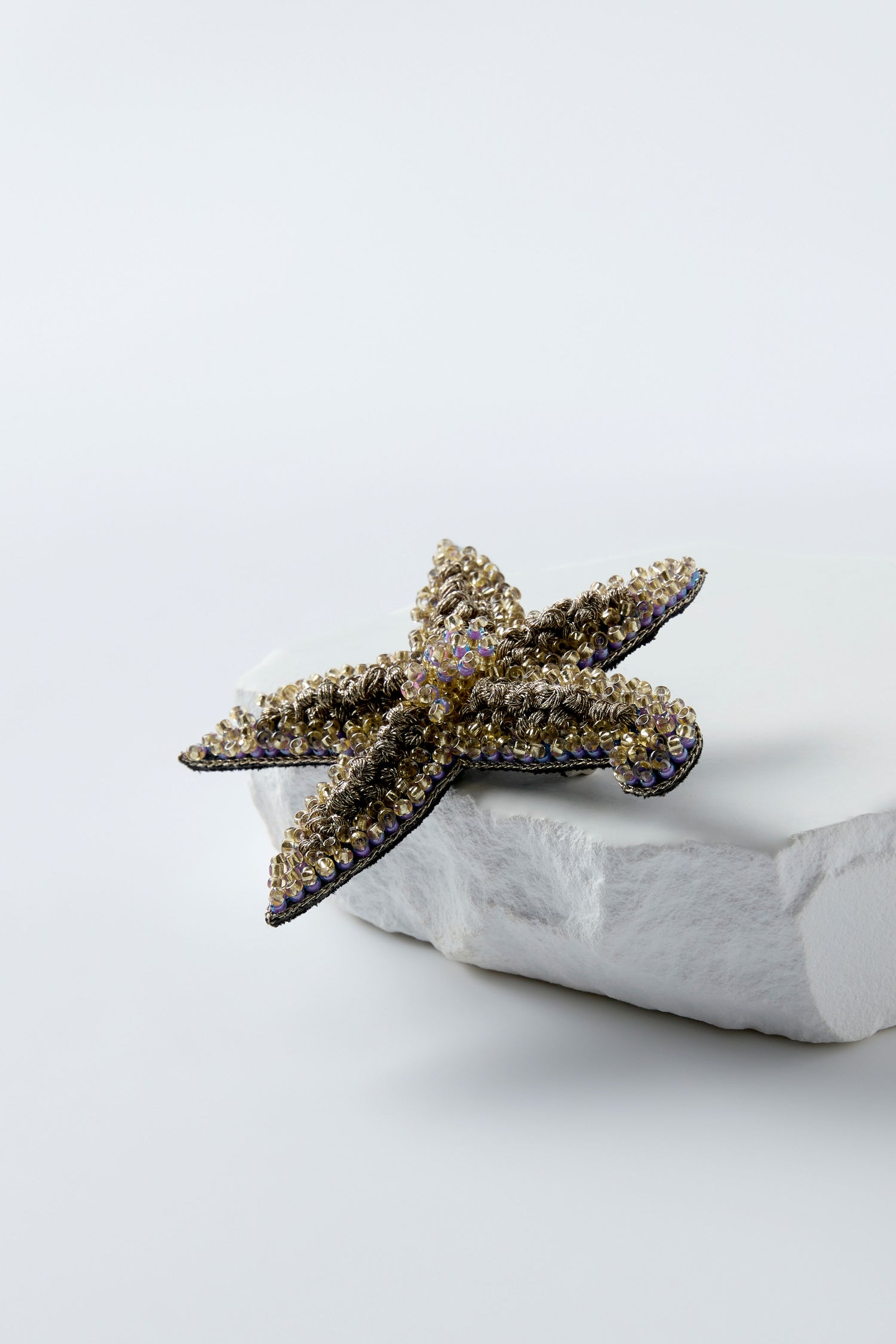 The Hand Beaded Starfish Brooch, crafted with gold and purple crystal beads, is displayed on a white stone platform against a plain background.