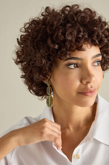 A person with curly hair wearing Hand Embroidered Crystal Bead Earrings in vibrant colors and a white shirt looks sideways, gently touching their collar.