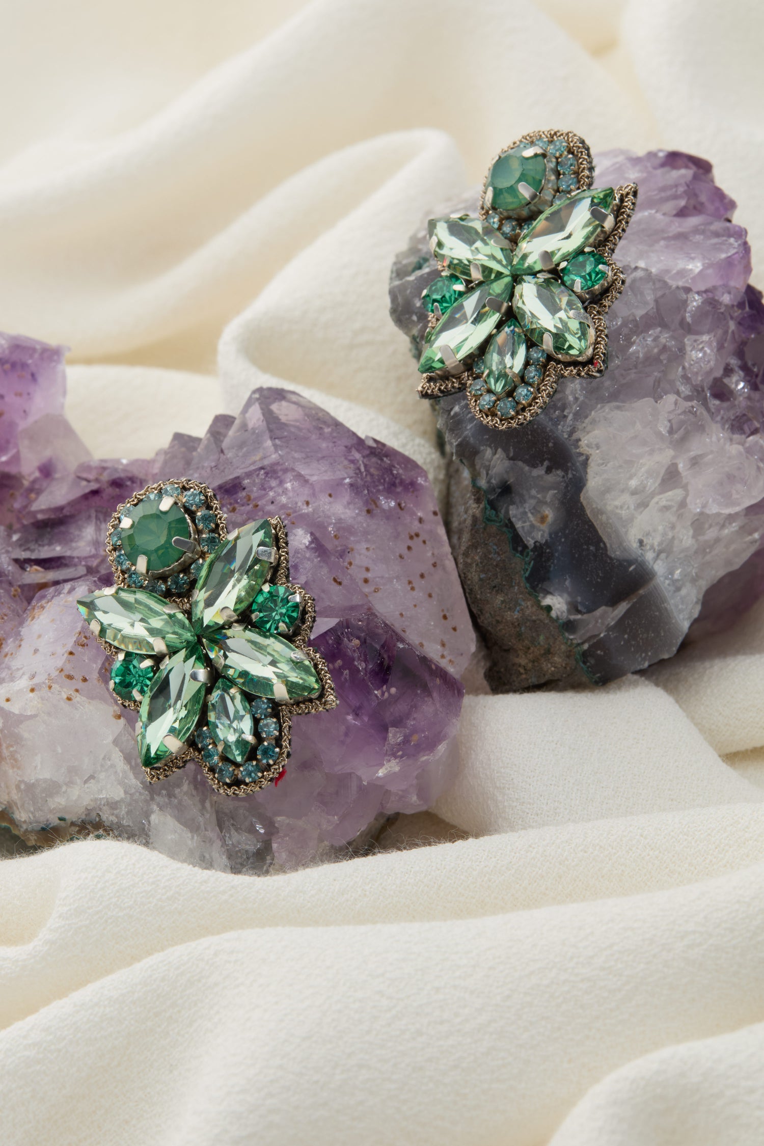 Two ornate, green jeweled brooches resembling butterflies are displayed on purple stones, set against a soft, light-colored fabric background. Their design mirrors the intricately embroidered details found on Hand Beaded Petal Earrings.