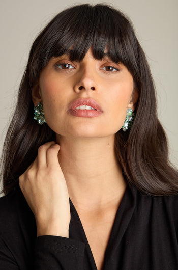 A woman with long dark hair and bangs is wearing Hand Beaded Petal Earrings in emerald green and a black top. She is looking straight into the camera with one hand touching her neck.