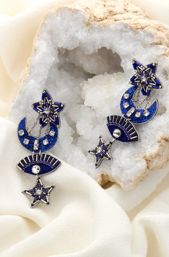 A pair of Celestial Hand Beaded Earrings featuring blue and silver celestial elements such as stars, moons, and eyes, displayed on a white crystal rock. These earrings embody the night sky's essence, making them a stunning piece of artisan jewelry.