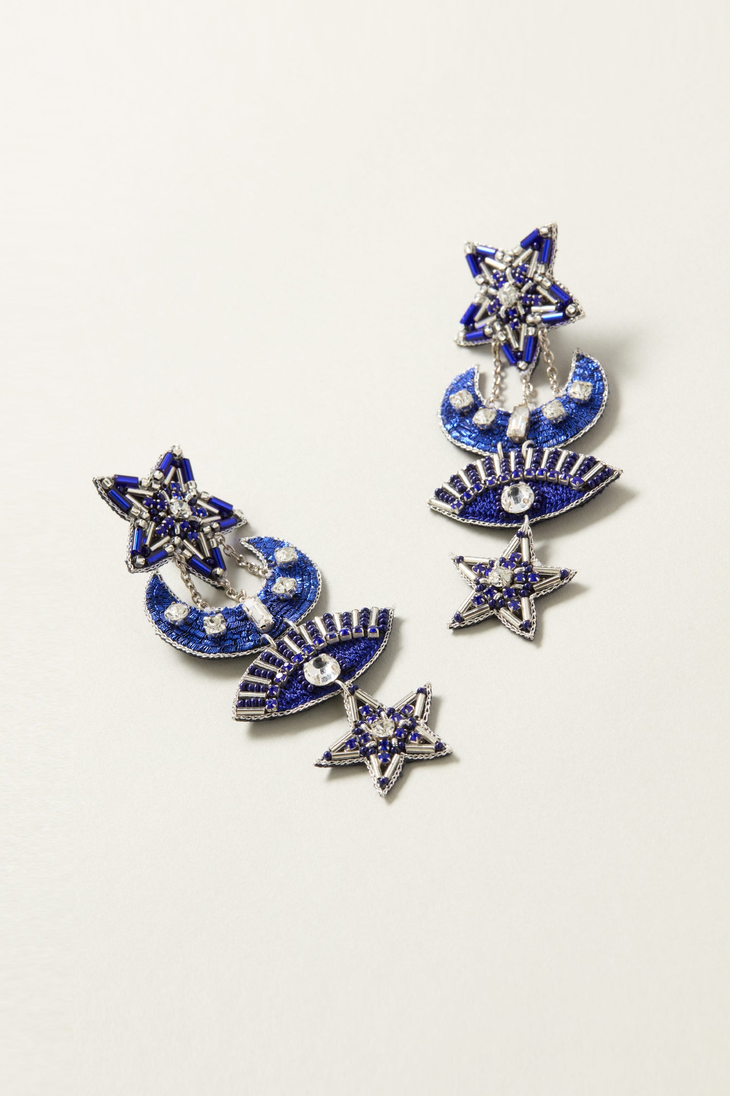 A pair of Celestial Hand Beaded Earrings, ornately designed in blue and silver with stars, crescent moons, and eye motifs embellished with small gemstones reminiscent of the night sky, arranged side by side on a white surface.