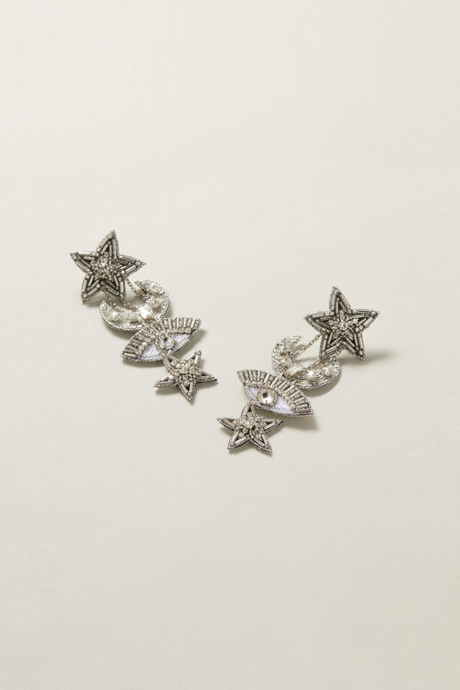 A pair of silver earrings adorned with stars and crescent moons, embedded with small, clear gemstones that evoke the night sky, arranged symmetrically on a plain, light background. These Celestial Hand Beaded Earrings are a true piece of artisan jewellery.