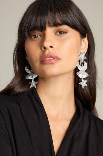 A woman with long dark hair and bangs wears large, ornate Celestial Hand Beaded Earrings featuring star and geometric designs. She has a neutral expression and is dressed in a black top, evoking the mysterious allure of the night sky.