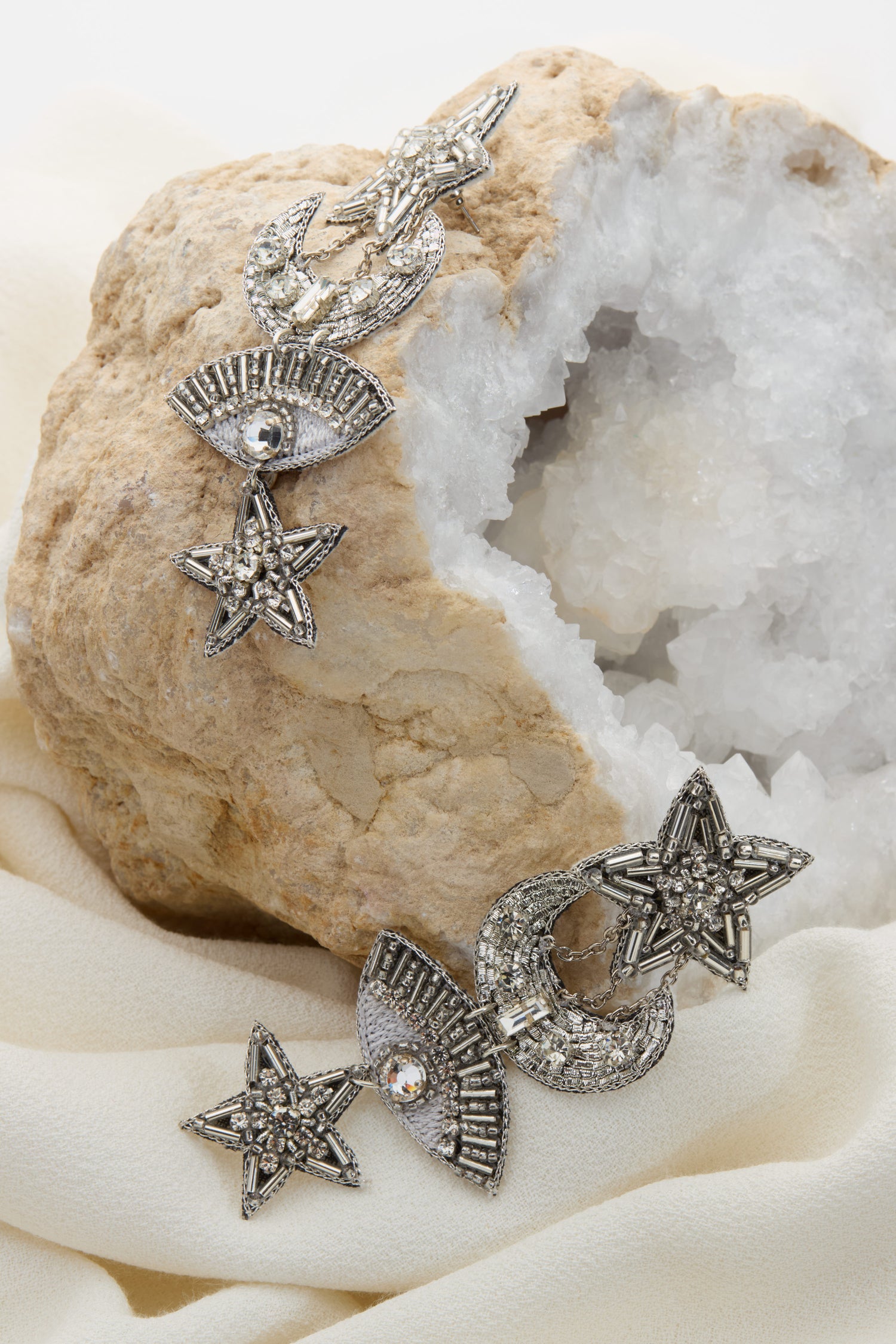 A pair of Celestial Hand Beaded Earrings, crafted in silver with intricate star-shaped designs and additional geometric motifs, is showcased on a piece of hollow rock with a crystalline interior. They capture the beauty of the night sky and exemplify true artisan jewelry craftsmanship.