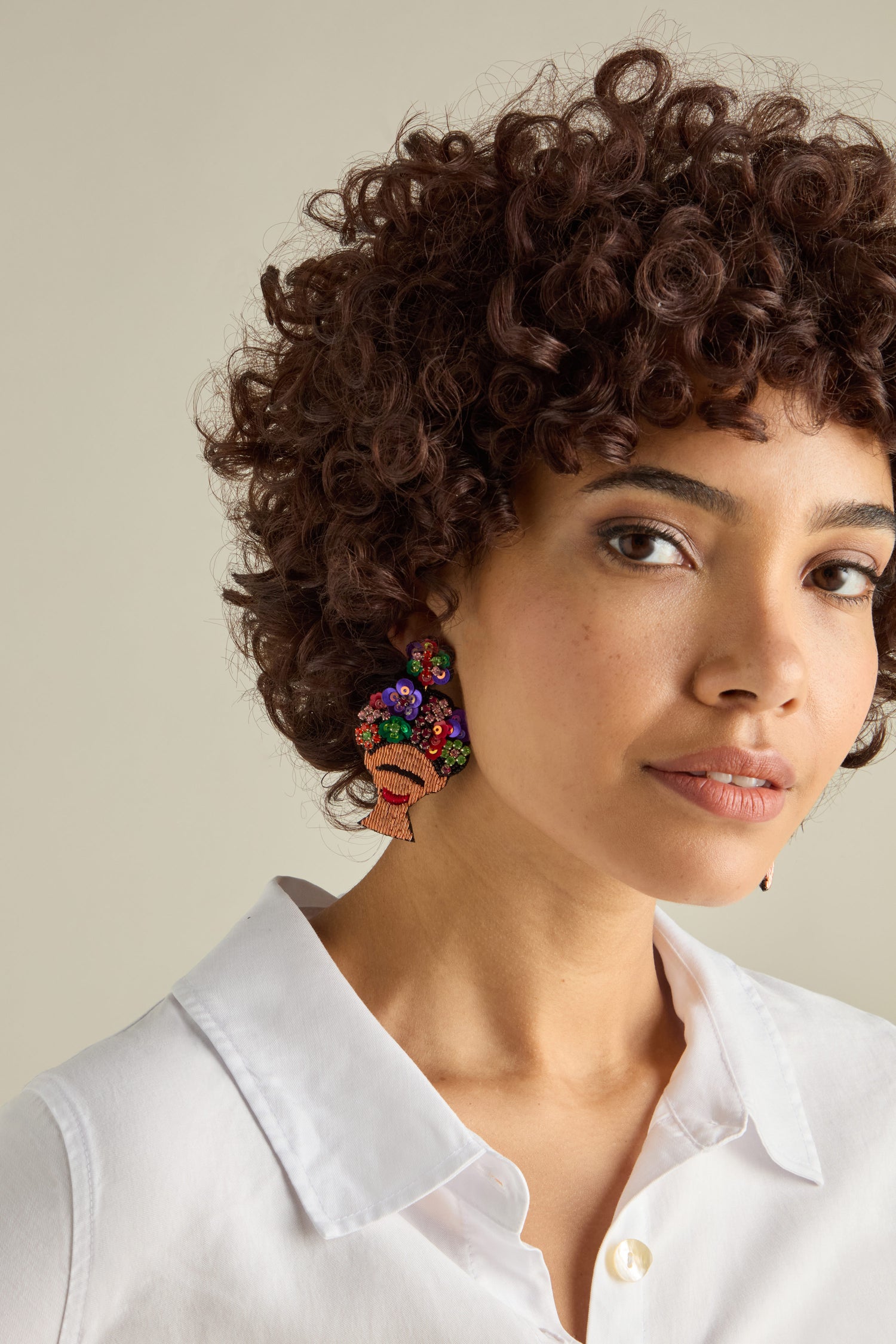 A person with curly hair in a white shirt wears stunning Hand Beaded Frida Earrings, shaped like a head with an afro.