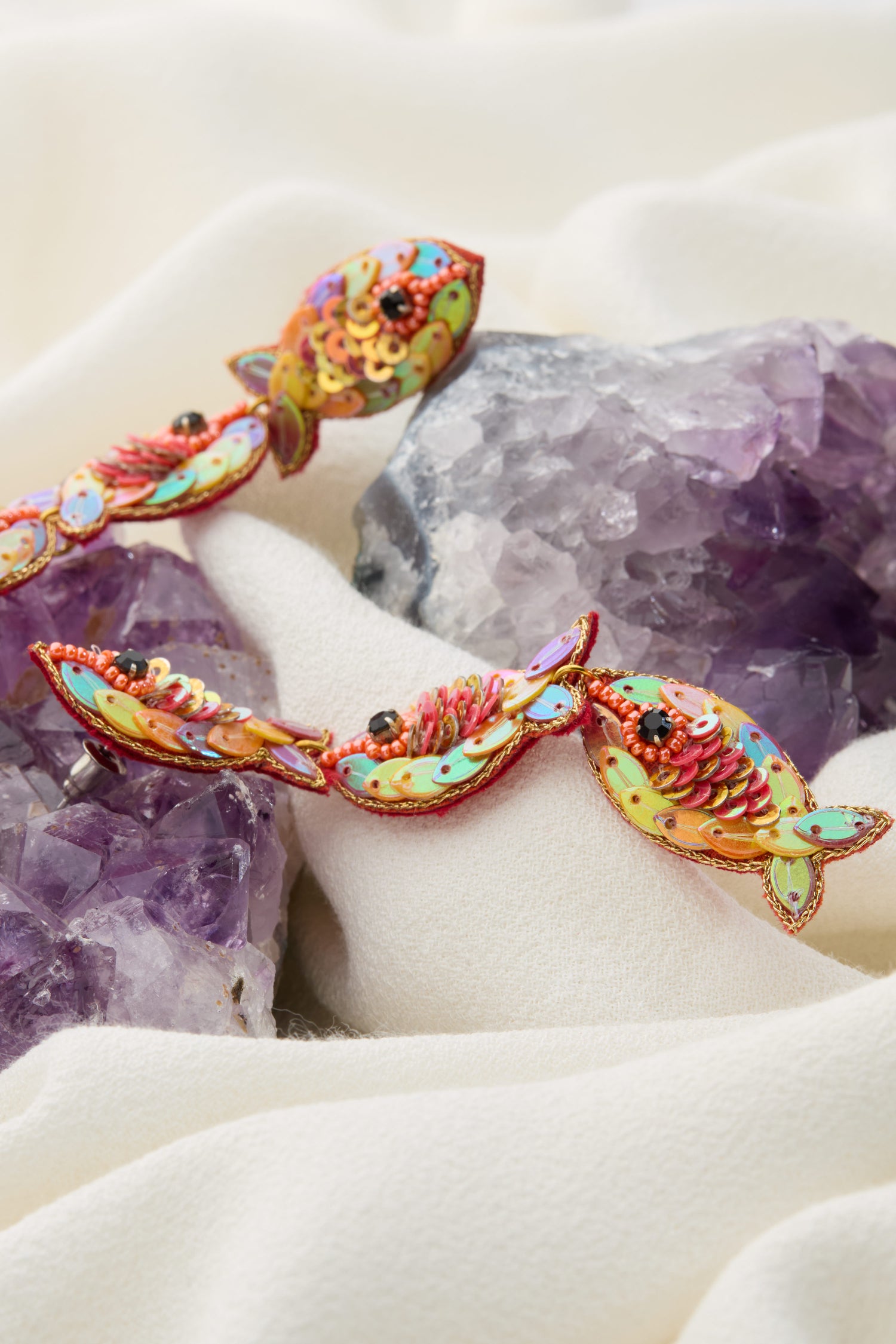 The Hand Beaded Poisson Earrings, featuring vibrant iridescent beads and sequins, rest on elegant purple crystals and white fabric, encapsulating the whimsical beauty of the ocean.