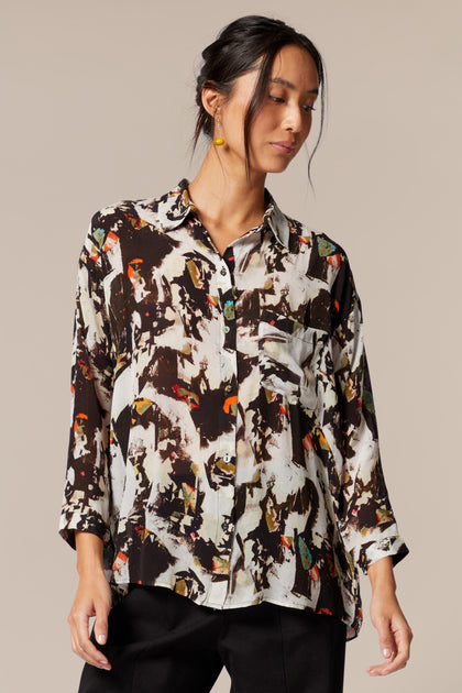 A model wears a shirt with bracelet-length sleeves, collar, and an abstract print in primarily brown and white with orange and green accents. The dress is worn with black trousers. 