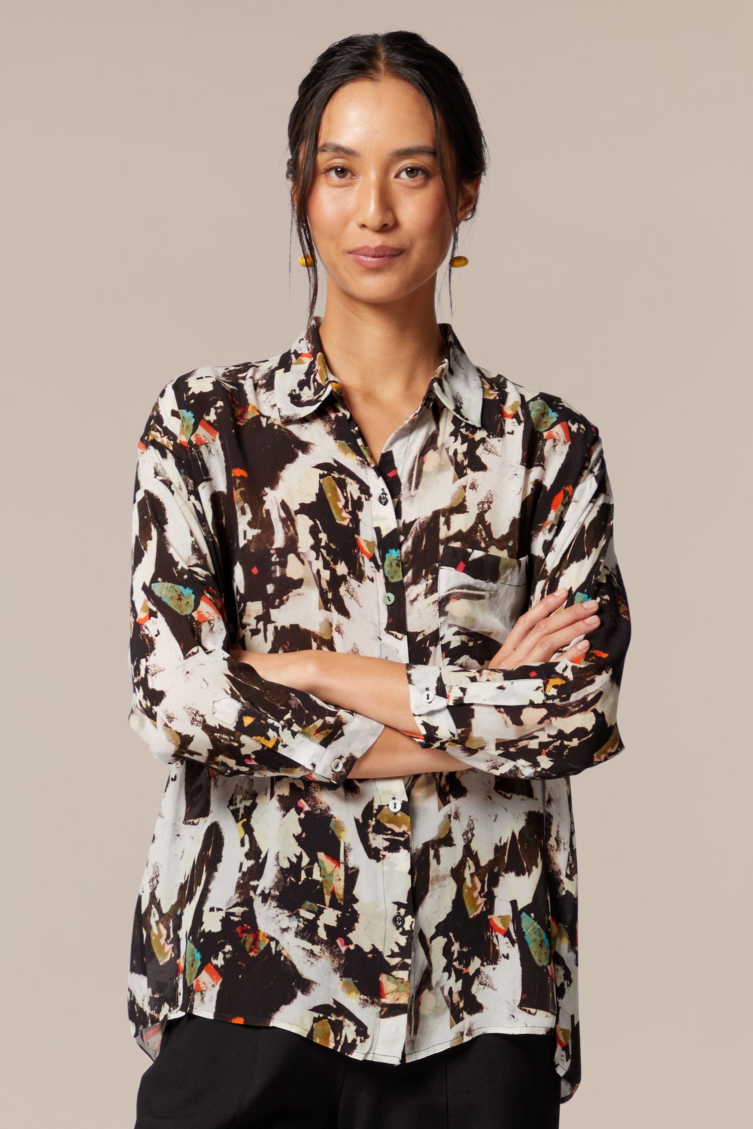 A model wears a shirt with bracelet-length sleeves, collar, and an abstract print in primarily brown and white with orange and green accents. The dress is worn with black trousers. 