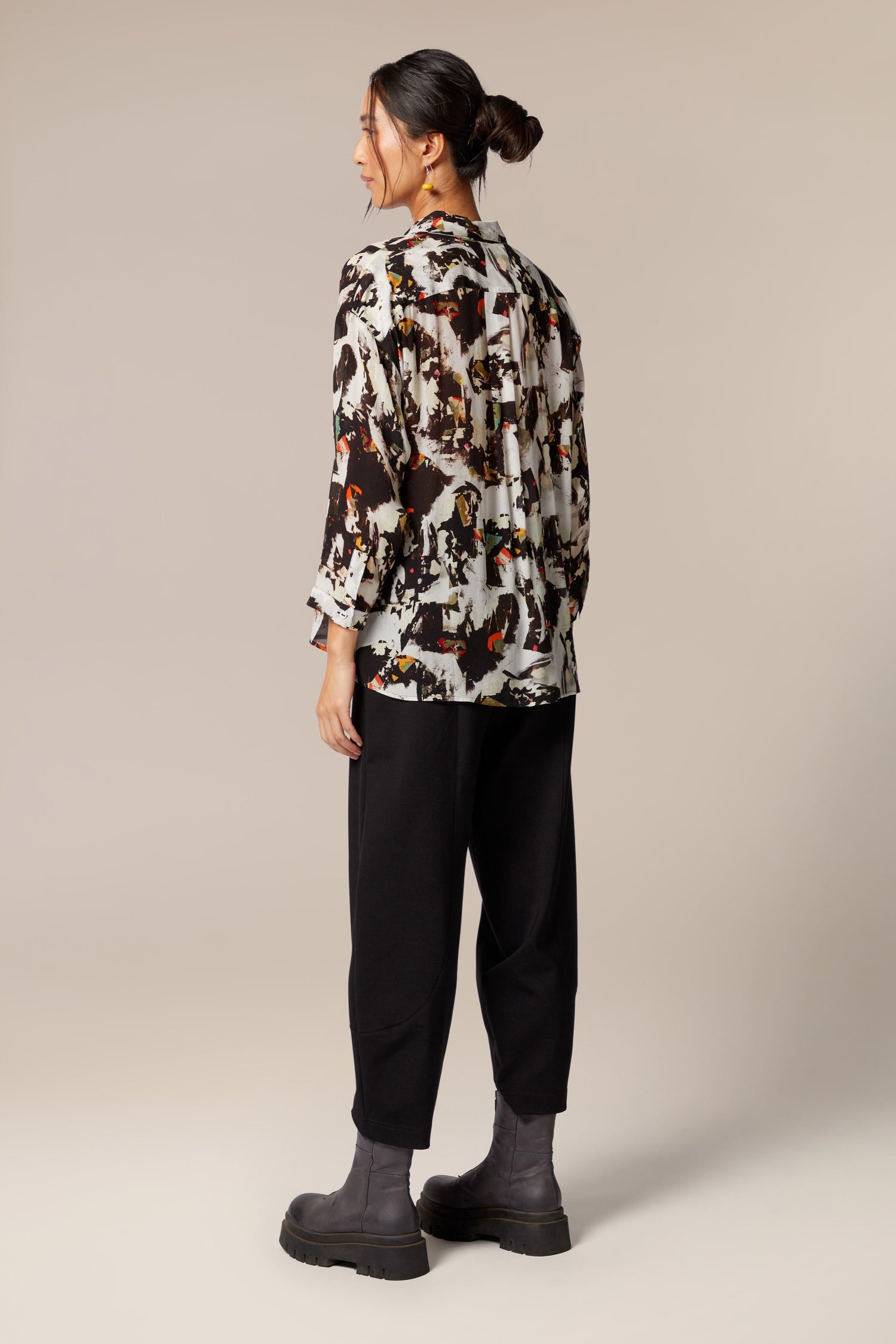 A model wears a shirt with bracelet-length sleeves, collar, and an abstract print in primarily brown and white with orange and green accents. The dress is worn with black trousers and leather zip boots.