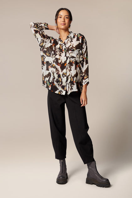 A model wears a shirt with bracelet-length sleeves, collar, and an abstract print in primarily brown and white with orange and green accents. The dress is worn with black trousers and leather zip boots.