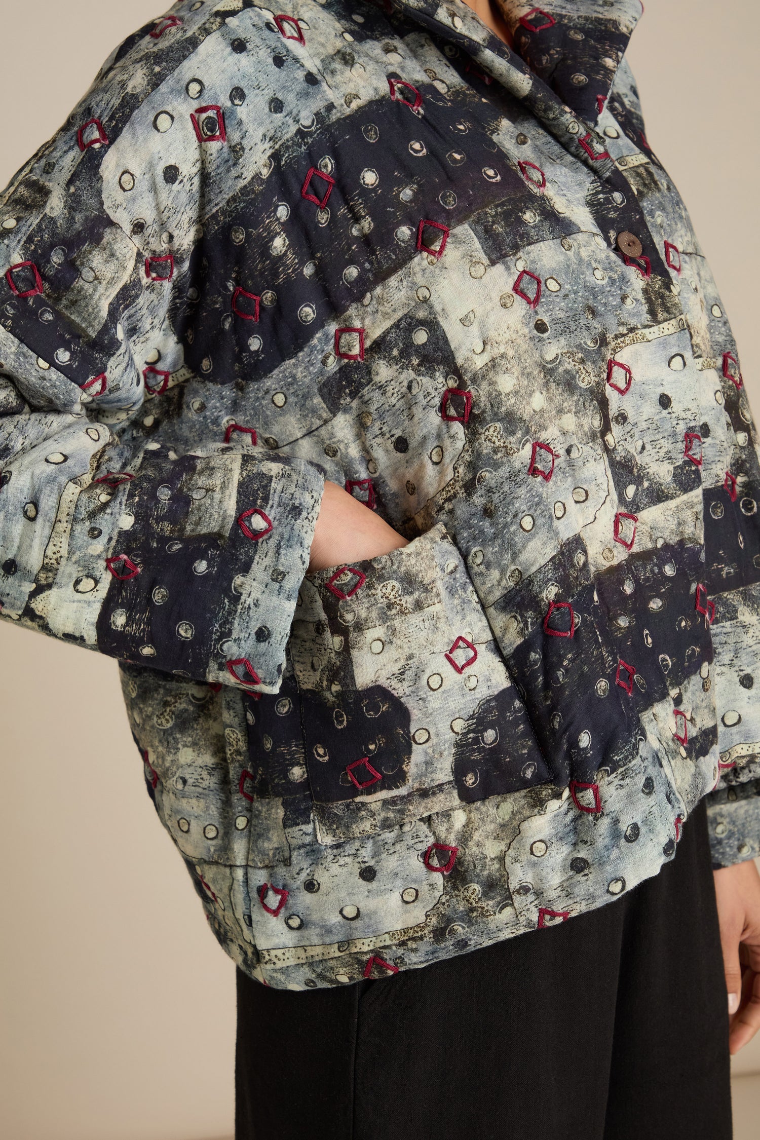 Close-up of a person wearing a Padded Embroidered Cotton Jacket with a geometric pattern of squares and circles in shades of blue and red, with detailed embroidery and their hand placed in the front pocket.