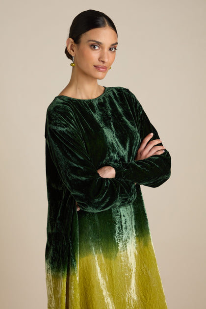 A person with dark hair stands with arms crossed, wearing a Dip Dye Silk Velvet Dress that fades to yellow at the bottom, showcasing a dip dye ombre effect.