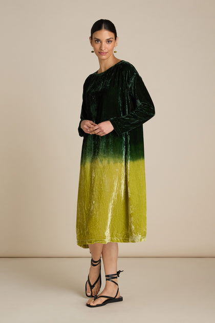 A person stands wearing an elegant Dip Dye Silk Velvet Dress with long sleeves, showcasing an ombre effect, paired with black strappy sandals against a plain beige background.