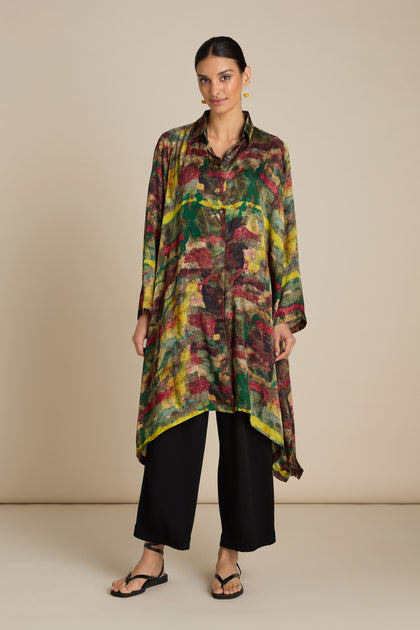 A person stands against a plain background wearing an Abstract Camo Shirt Dress over black pants and black sandals. The shirt dress has an asymmetrical hem and is paired with gold hoop earrings.