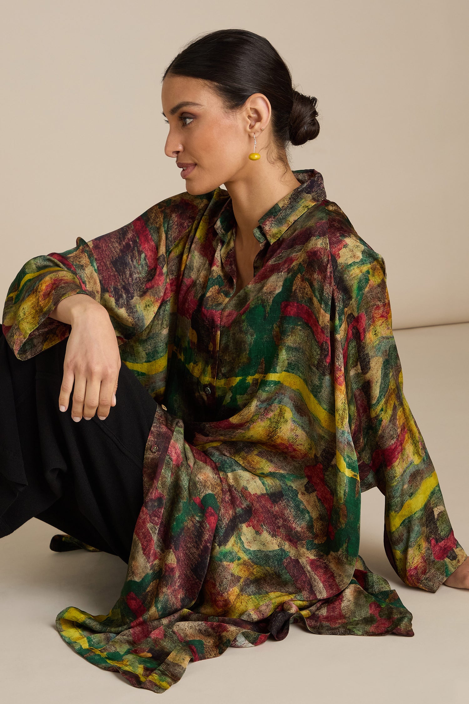 A person with dark hair in a bun is seated on the floor, wearing an Abstract Camo Shirt Dress and black pants.