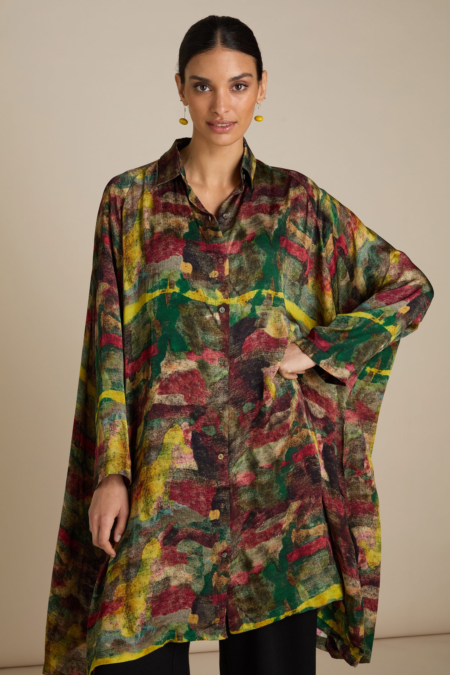 A woman stands wearing the Abstract Camo Shirt Dress, a colorful, loose-fitting, button-up shirt with a mix of green, yellow, red, and black hues. Reminiscent of painterly inspired prints, the silky viscose fabric adds an elegant touch. She has her right hand on her hip and is standing against a plain background.