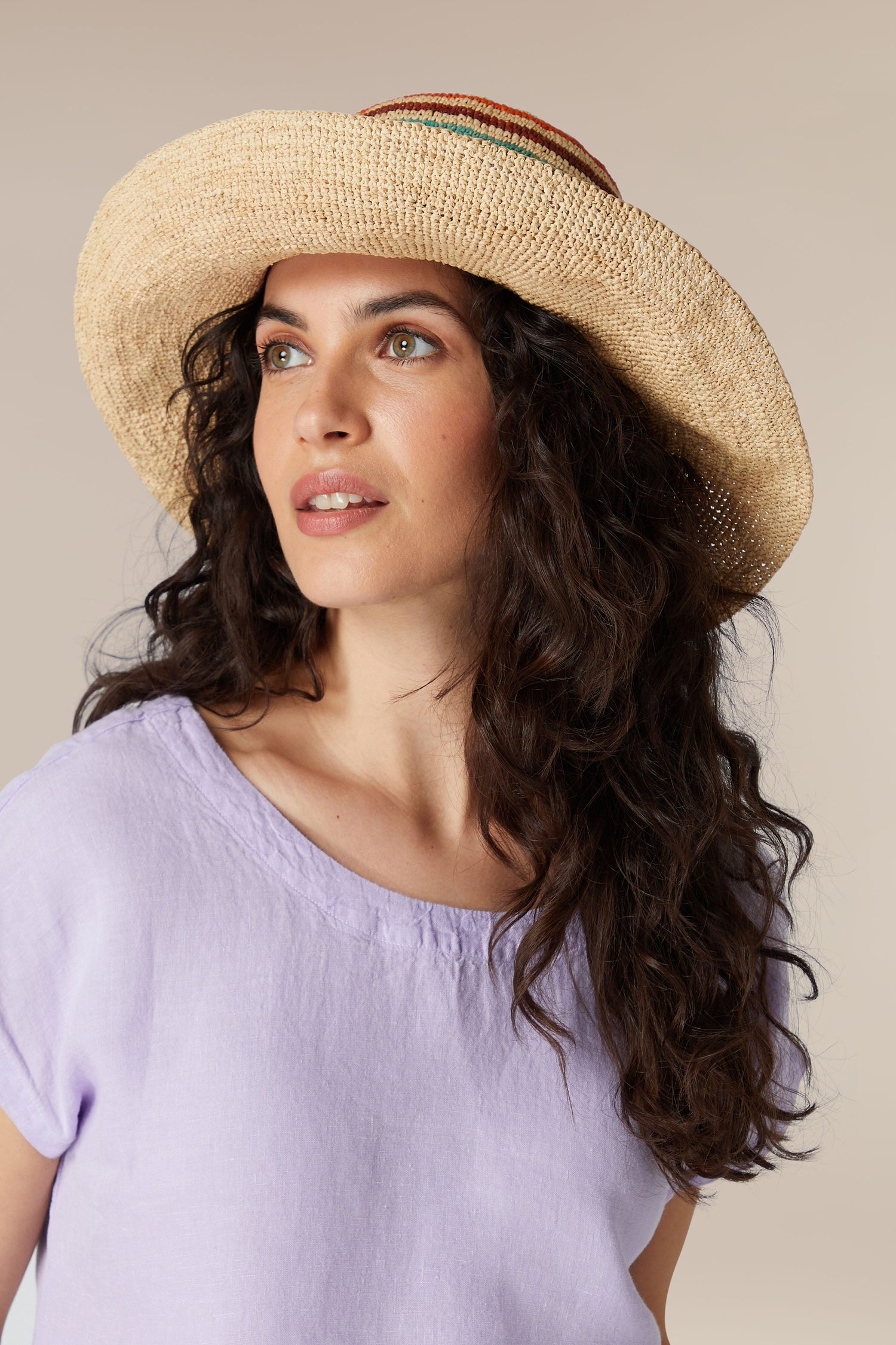 A woman with wavy brown hair wearing a Straw Boater Hat and a lilac top looking away from the camera.