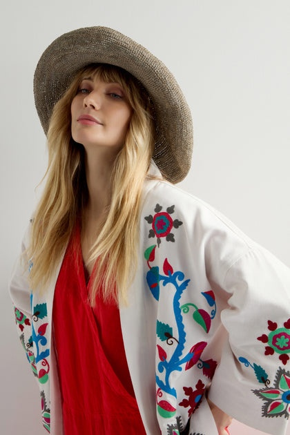 A woman in a vibrant red dress and an embroidered white jacket stands gracefully against a plain background, protected from the sun by a Handwoven Wide Brim Raffia Hat.