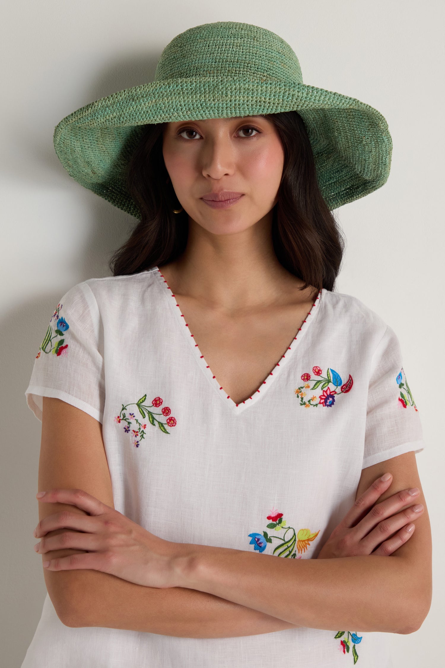 A woman wearing the Handwoven Wide Brim Raffia Hat and a white V-neck blouse with colorful floral embroidery stands with her arms crossed against a plain background.
