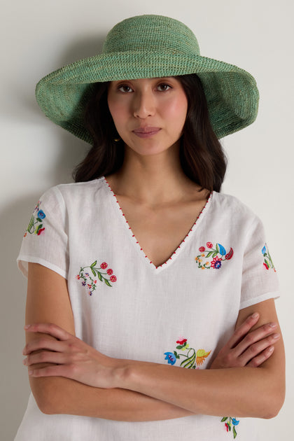 A woman wearing the Handwoven Wide Brim Raffia Hat and a white V-neck blouse with colorful floral embroidery stands with her arms crossed against a plain background.