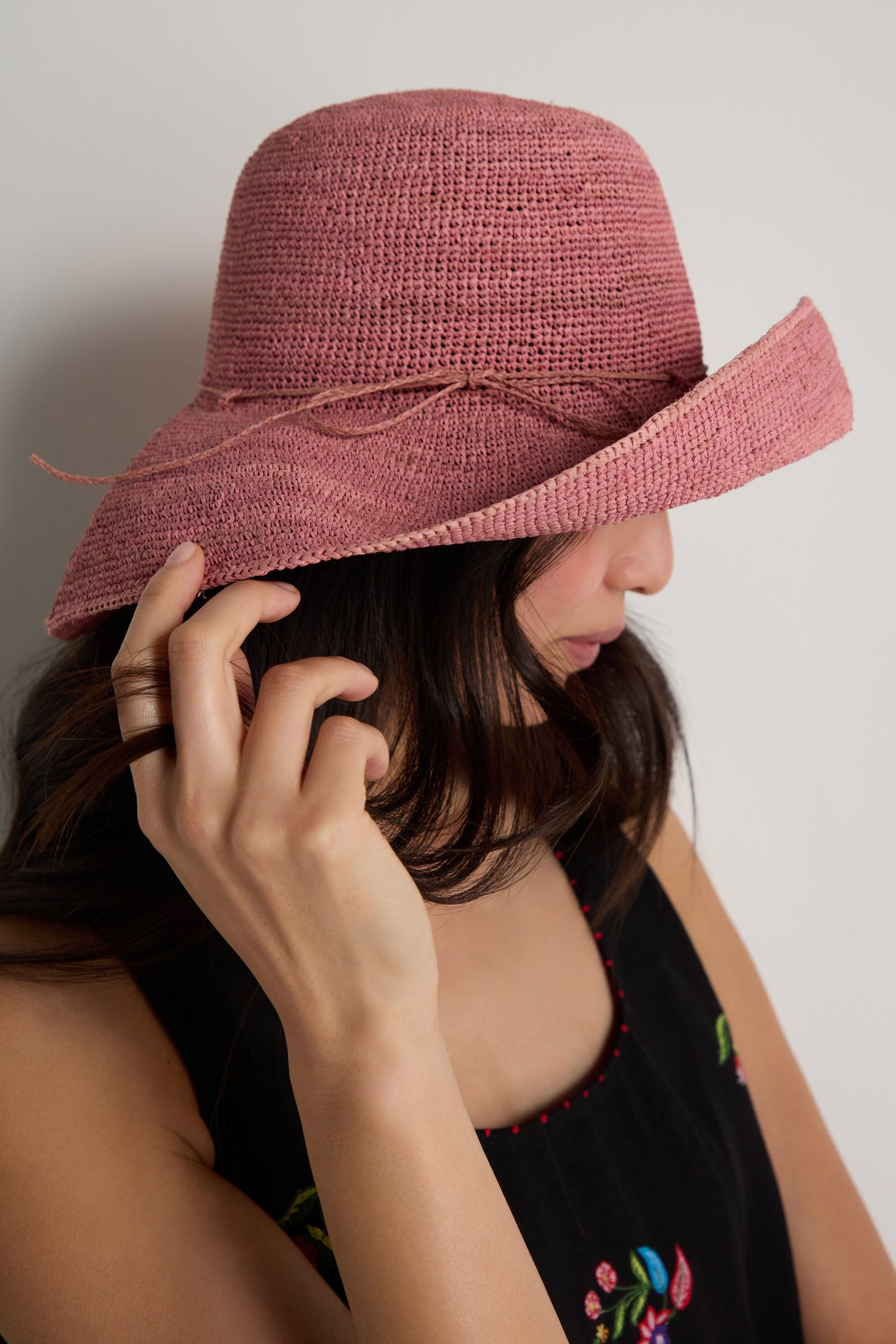 A woman wearing the Handwoven Wide Brim Raffia Hat in pink and a black sleeveless floral top stands sideways, gently touching the brim of her hat.