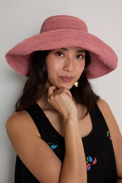 A woman poses against a plain background, wearing a black sleeveless top and the Handwoven Wide Brim Raffia Hat in pink, with one hand resting thoughtfully under her chin.