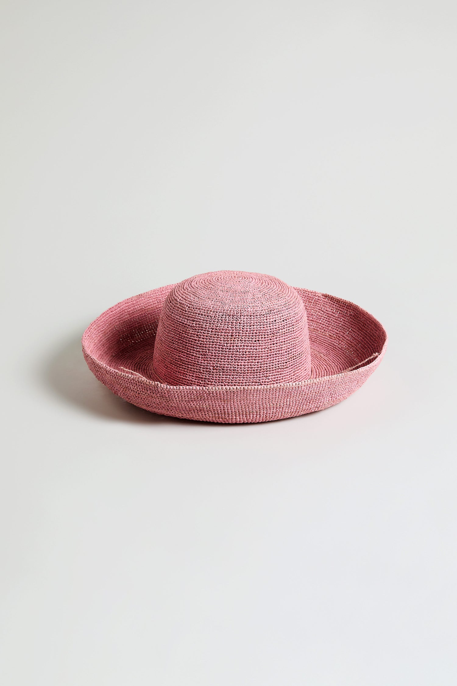 The Handwoven Wide Brim Raffia Hat in pink, crafted in Madagascar with a wide, slightly upturned brim, is displayed against a plain light background.