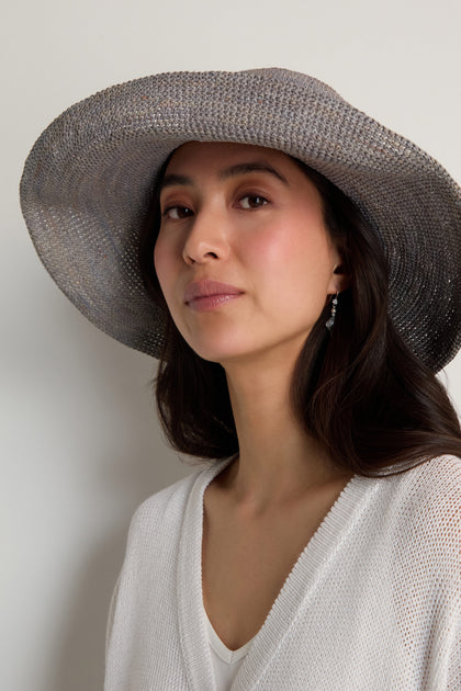 A woman stands against a plain light background wearing the Handwoven Wide Brim Raffia Hat, paired with a white knit top and drop earrings.