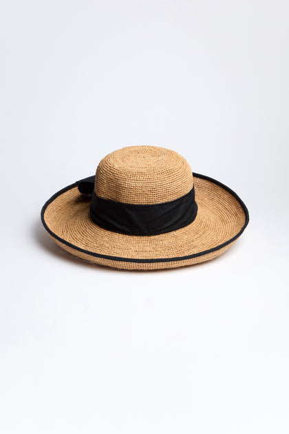 The Handwoven Ribbon Raffia Hat from Madagascar flaunts a wide brim and a black band on a white background. Ideal for sun protection, it seamlessly blends style and functionality.